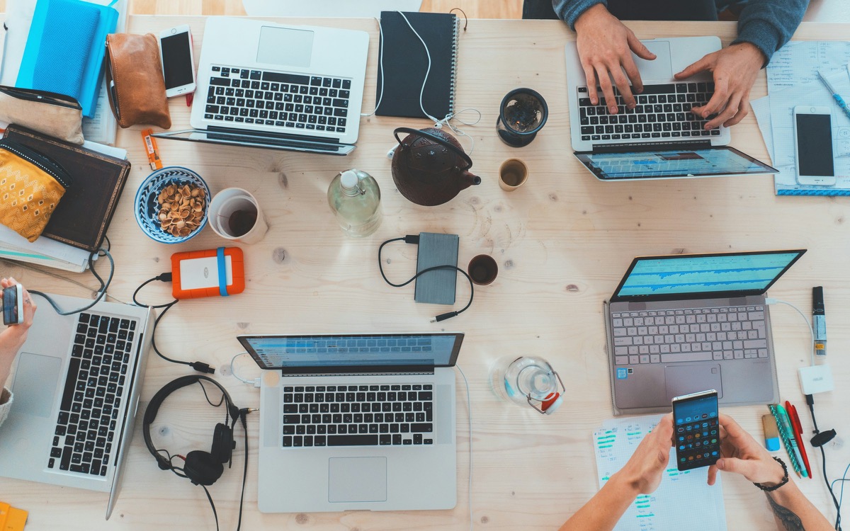Tavolo di lavoro in agenzia: più laptop, grafici e dispositivi — il collage di fine mese tra piattaforme